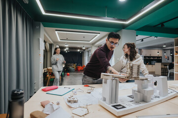 A dynamic group works on an architectural model at a large table in a modern collaboration center, with others in the background. The scene conveys teamwork, creativity, and hands-on learning.