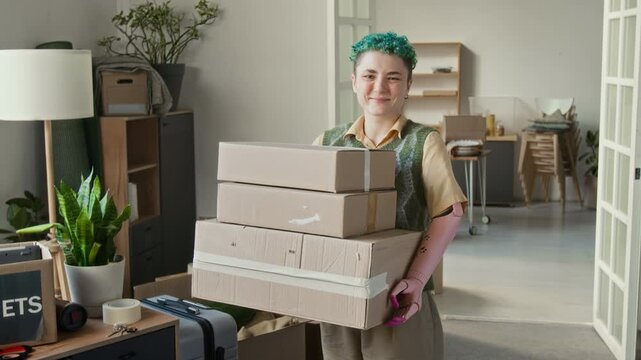 Portrait of Caucasian short-haired girl with prosthetic arm smiling for camera holding three cardboard boxes while relocating to new apartment