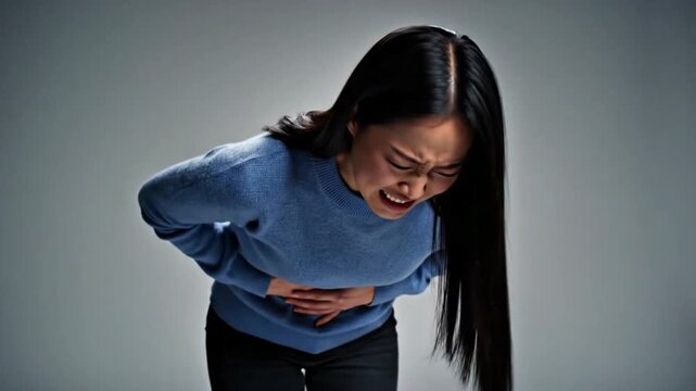 Asian Woman in Blue Sweater Holding Stomach in Severe Pain on Gray Background