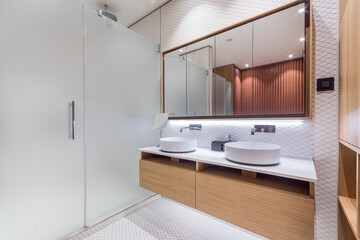 This bathroom features two round sinks on a wooden vanity. A large mirror reflects the light. A clear glass shower is visible along with tiled flooring and wooden accents.