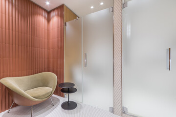 This setting features a contemporary bathroom design with a unique chair and a small table. Frosted glass panels separate the shower area, creating a clean look. Soft lighting enhances the space.