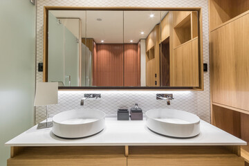 The bathroom features a countertop with two round sinks, a large mirror, wooden cabinets, and soft lighting. The space is neat and organized, showing clear design lines.