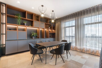 A dining area features a large table and black chairs. Shelves are filled with plants and other simple items. Natural light enters through large windows during the day.