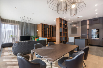 The scene shows a modern interior space that includes a dining area with a large table and dark chairs. A living room is visible in the background with a sofa and a TV. Large windows let in light. © Alhim