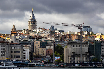 Fototapeta premium The historic Galata Tower contrasts with a construction crane on the Karakoy waterfront under dramatic cloudy skies lit by side sunlight. A scene of Istanbul's constant evolution. Istanbul, Turkey.