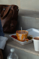 Informal lunch of corn on the cob in a cup with cheese and messy overflowing jalapeño peppers in a white polystyrene container on cement steps next to a travel bag, fast food to go.