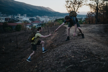 Two backpackers on a steep hillside trail assist a partner as they traverse a rough incline, capturing teamwork, endurance, and outdoor adventure against a backdrop of a distant town and mountains.