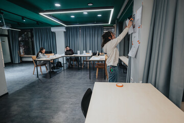 A contemporary classroom scene with students at desks and a team member attaching notes to a wall, reflecting collaboration, learning, and a focused study atmosphere in a shared space.