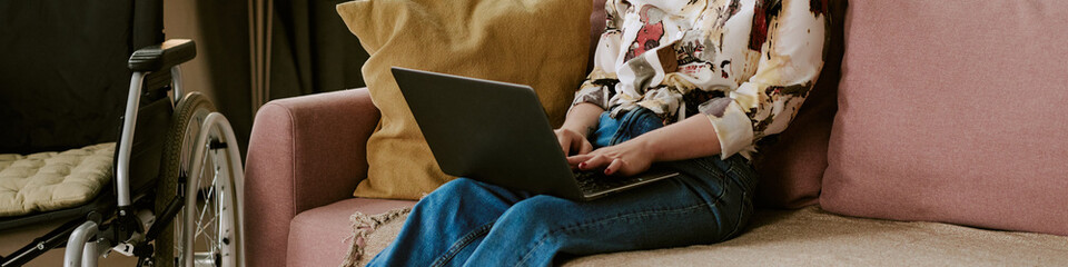 Website banner of woman with disability sitting on sofa using laptop, wheelchair visible beside...