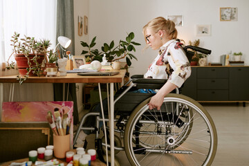 Caucasian young adult woman with disability sitting in wheelchair working at desk surrounded by plants and art supplies, focusing on creative project in home workspace