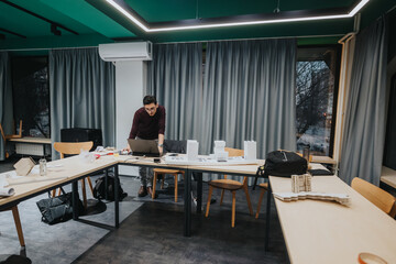 A student collaborates at a bright collaboration center, using a laptop while reviewing architectural models and materials on long tables. Modern lighting and curtains create a focused, productive