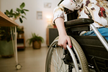 Caucasian young adult woman with disability sitting in wheelchair indoors, hand gripping wheel, partial body visible, demonstrating independence and mobility in daily life
