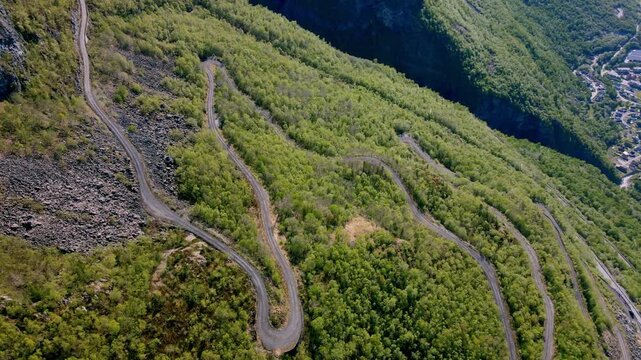 Aerial drone view of a winding mountain road with sharp hairpin turns and switchbacks climbing a steep green slope in the Norwegian wilderness.