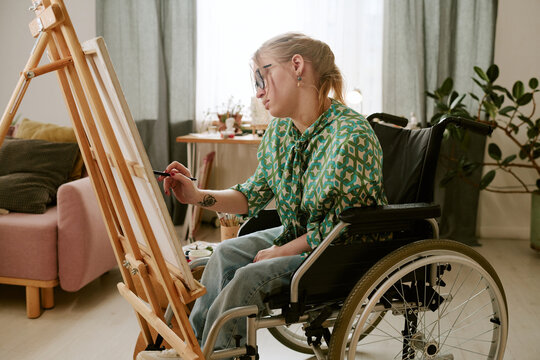 Caucasian young adult woman with disability sitting in wheelchair painting on canvas using brush, focused on creative process in bright room with natural light and plants visible - Powered by Adobe
