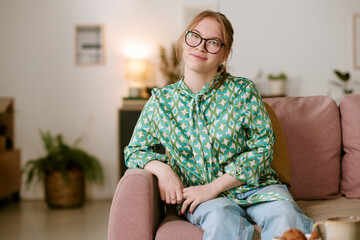 Fototapeta premium Portrait of Caucasian woman with blond hair and glasses sitting on sofa smiling at camera, hands resting on armrest, relaxed posture in modern living room setting
