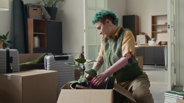 Short-haired gen Z girl with pink bionic arm unpacking cardboard box after moving into new apartment with her boyfriend
