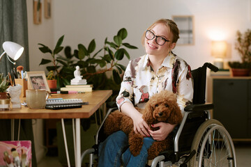 Portrait of young adult Caucasian woman with disability sitting in wheelchair smiling at camera holding brown poodle dog in home office workspace