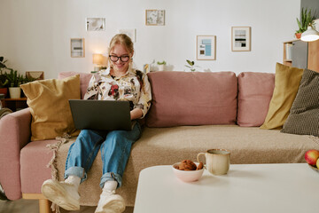 Teenage Caucasian girl sitting on sofa using laptop, smiling while looking at screen, wearing glasses, relaxing in modern living room