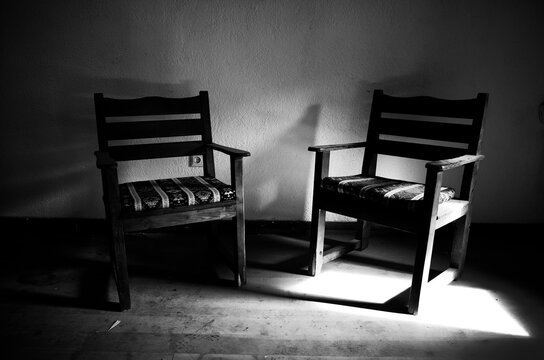 Two empty wooden chairs in a dark room with a patch of sunlight on the floor, concept of loneliness and absence