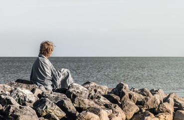 A small boy sits alone on boulders wearing a beach towel. He gazes over the sea in Norway, enjoying the summer day and taking in the view with no one around him