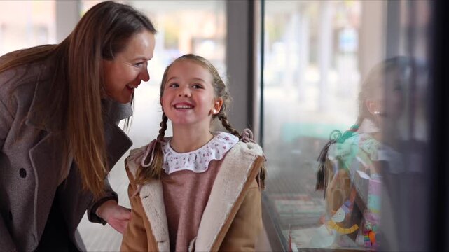 Adorable little girl talking and smiling with her loving mother while shopping together for gifts, their happy reflections visible in the store window on a beautiful sunny day