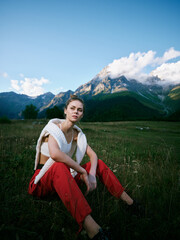 Naklejka premium Woman sitting on a meadow grass in redpants and a light knit top against a vast mountain landscape, clear sky and open fields, showcasing outdoor fashion, calm mood, and nature inspired scenery
