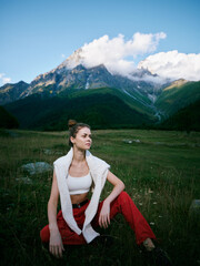 Naklejka premium Outdoor scene with a woman seated in a grassy meadow, mountains above and clouds in a clear blue sky, casual athletic wear, relaxed pose, nature and solitude in natural light and open space