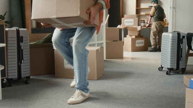 Gen Z man stumbling over cardboard box labeled books when carrying another one into room, he placing it down and rubbing hit leg