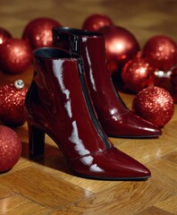 Pair of glossy deep red ankle boots with a zipper surrounded by red Christmas baubles. Festive and elegant footwear for winter celebrations.