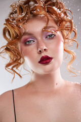 model stands in front of a light background, showing off her curly red hair styled with snow-like...