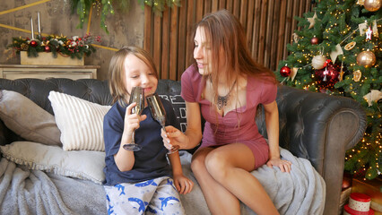 A beautiful young woman clinks a festive glass with a funny long-haired boy on a sofa near a Christmas tree