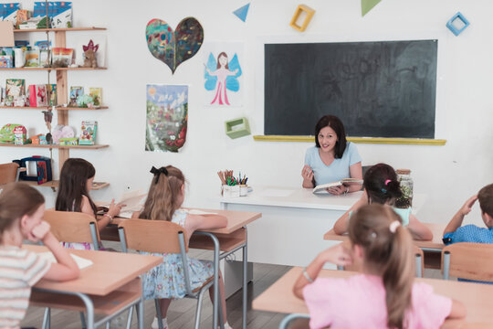 A teacher reads a book to elementary school students who listen carefully while sitting in a modern classroom - Powered by Adobe