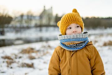 Smiling child in mustard jacket and knit hat crouches in snow, cheeks rosy from the cold. Winter...