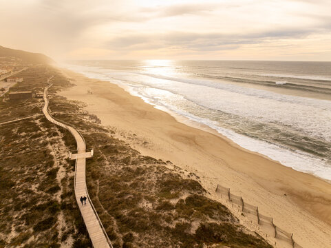 Aerial view of golden sands meet the foamy waves, a wooden path winds through dunes under a soft, hazy sky, Praia de Quiaios, Figueira da Foz, Portugal. - Powered by Adobe