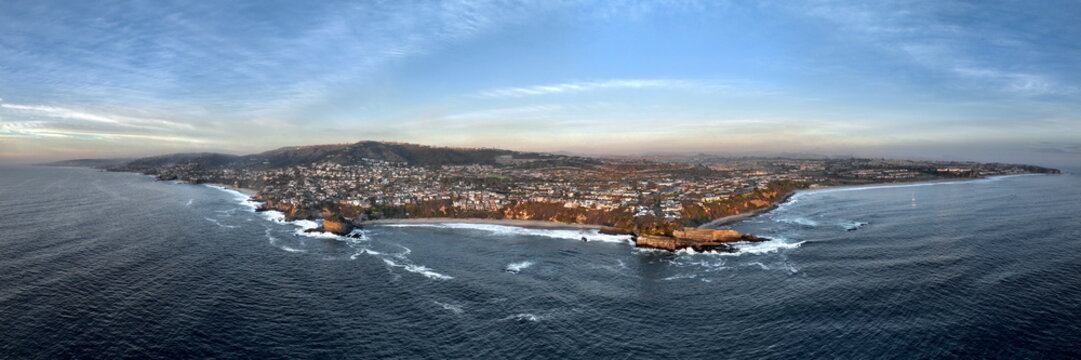 Aerial view of where the deep blue ocean meets the sandy beach and rocky cliffs under a soft, pale sky, Laguna Beach, California, United States.