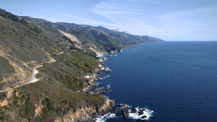 Aerial view of the winding coastal road snaking along the rugged cliffs, where the deep blue ocean crashes against the rocky shore, Big Sur, California, United States.