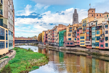 Vibrant Girona waterfront with colorful houses along the Onyar River, Gothic cathedral spire above the buildings, and autumn trees by the bridge on a cloudy day. Girona, Spain