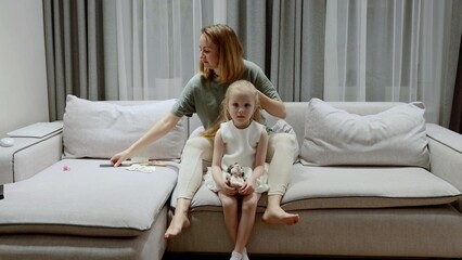 Mother gently brushing daughter's hair, sitting together on living room sofa, sharing intimate familial connection and tender care