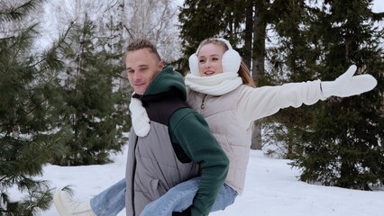Cheerful couple sharing piggyback ride through snow covered forest, wearing cozy winter attire, spreading arms playfully during festive holiday moment