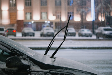 Car windshield and wipers layered in fresh snow with falling flakes, icy buildup and blurred city lights suggesting cold, challenging winter driving and parked vehicle prep