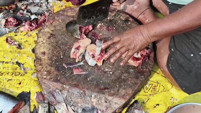 Maski, Karnataka, India &ndash; December 7, 2025: Vendor chopping fresh fish on a wooden block surrounded by fish parts and tools. A raw editorial look at traditional seafood preparation.
