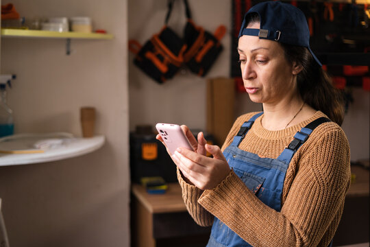 Portrait of female repairman or mechanic wearing overalls chatting online and shopping in internet using smartphone in garage workshop - Powered by Adobe