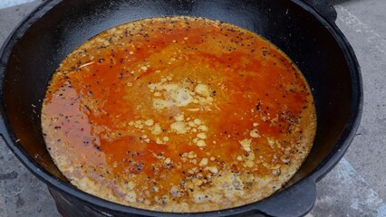 Heavy skillet cooks flavorful broth, Enormous iron vessel gently reduces fragrant stock, Vast metallic cooking vessel gently concentrates savory stock as vendor stirs amidst marketplace activity
