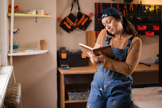 Portrait of female repairman or mechanic wearing overalls, craftsman multitasking writing notes phone call chat with client or customer in garage workshop. - Powered by Adobe