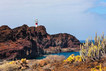 Rocky coastline of Tenerife shows bright summer landscape with Punta Teno lighthouse. Sunlit cliffs rise above blue ocean creating vivid coastal scenery with clear warm atmosphere.