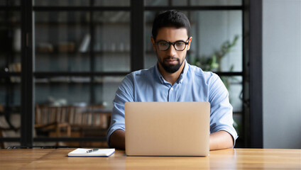 Serious young bearded indian man looking at laptop screen working online, focused male employee busy typing on computer keyboard sitting at desk in modern office, concentrated student learning