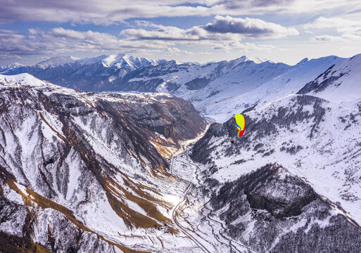 Aerial view of a paraglider soaring between snow-laden mountains and valleys under a sky streaked with clouds, Ganisi, Mtskheta-Mtianeti, Georgia.