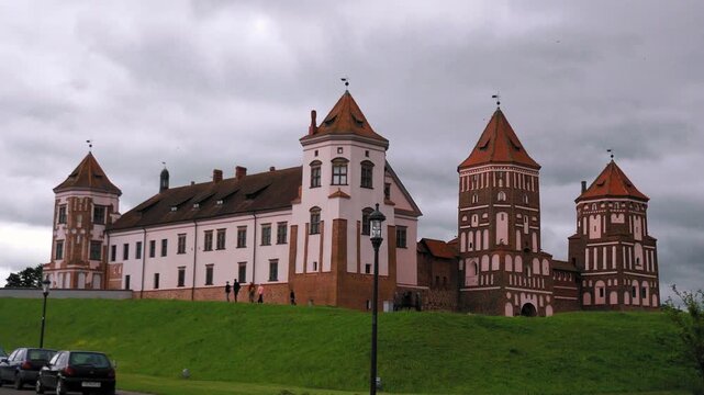 Majestic mir castle complex, a unesco world heritage site in belarus, standing on a green hill with people strolling along its walls under an overcast sky on a cloudy summer day