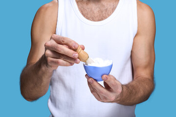 Caucasian young adult man holding shaving brush and bowl with shaving cream preparing for grooming routine against blue background, muscular arms and torso visible