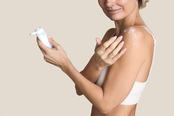 Caucasian middle aged woman applying moisturizing cream to shoulder while holding bottle, smiling slightly, showing upper body and hands, demonstrating skincare routine against plain background